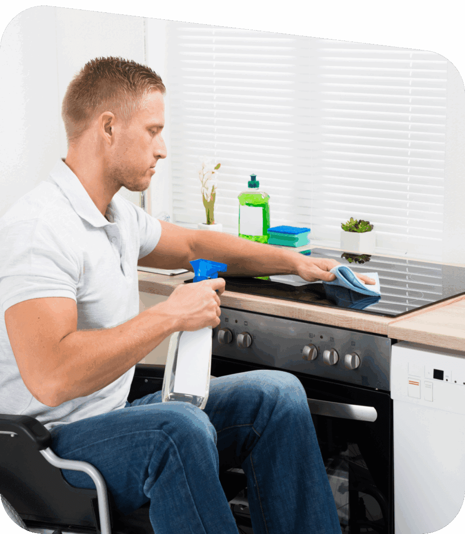 A man sitting in a wheelchair cleans a kitchen stovetop with a spray bottle and cloth, with cleaning supplies visible on the counter.