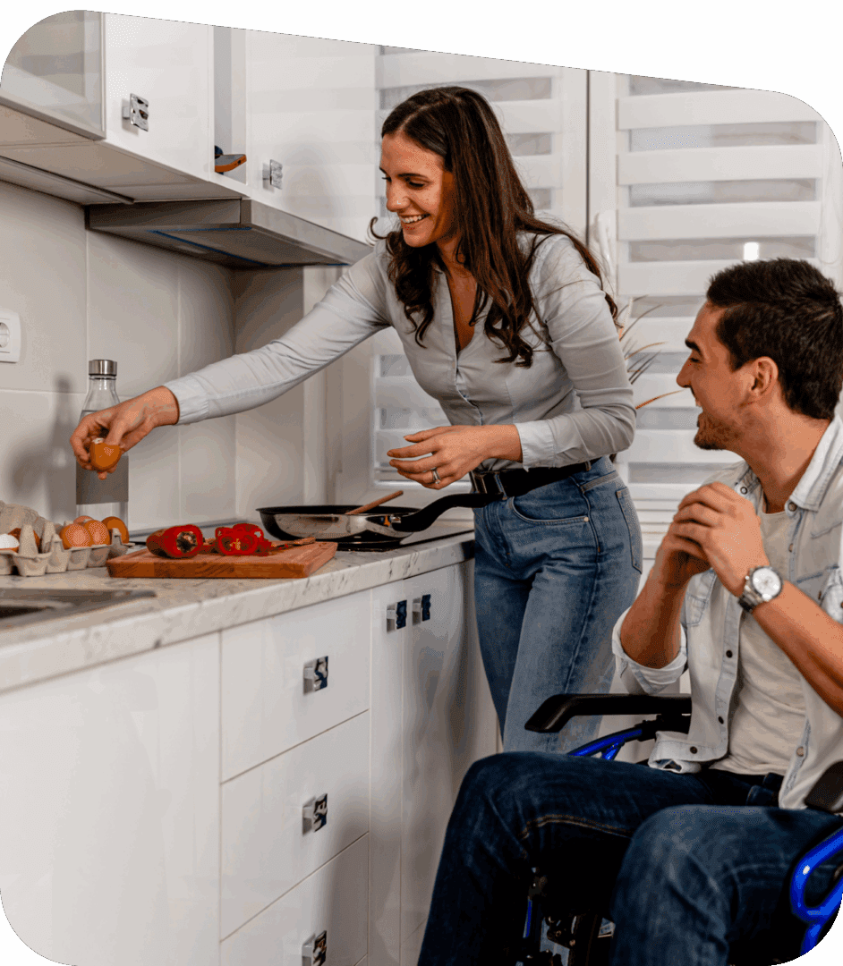 A woman prepares food at a kitchen counter while a man in a wheelchair watches and smiles.