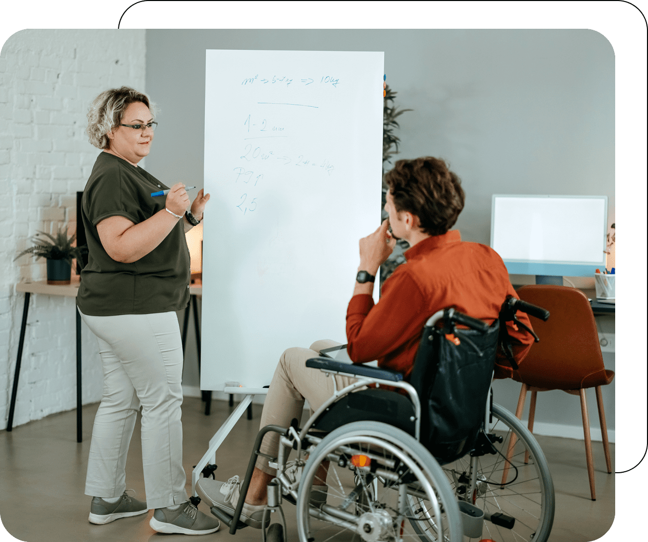 A woman writes on a whiteboard while a man in a wheelchair looks on in an office setting.