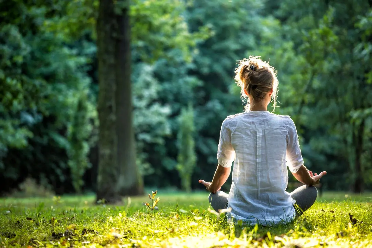 Person meditating outdoors on grass in a peaceful forest setting.
